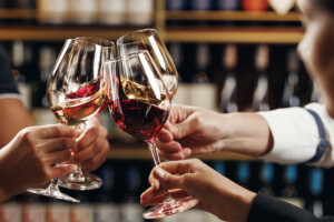 Photo of several wine glasses in a toast with shelves of wine bottles in the background.